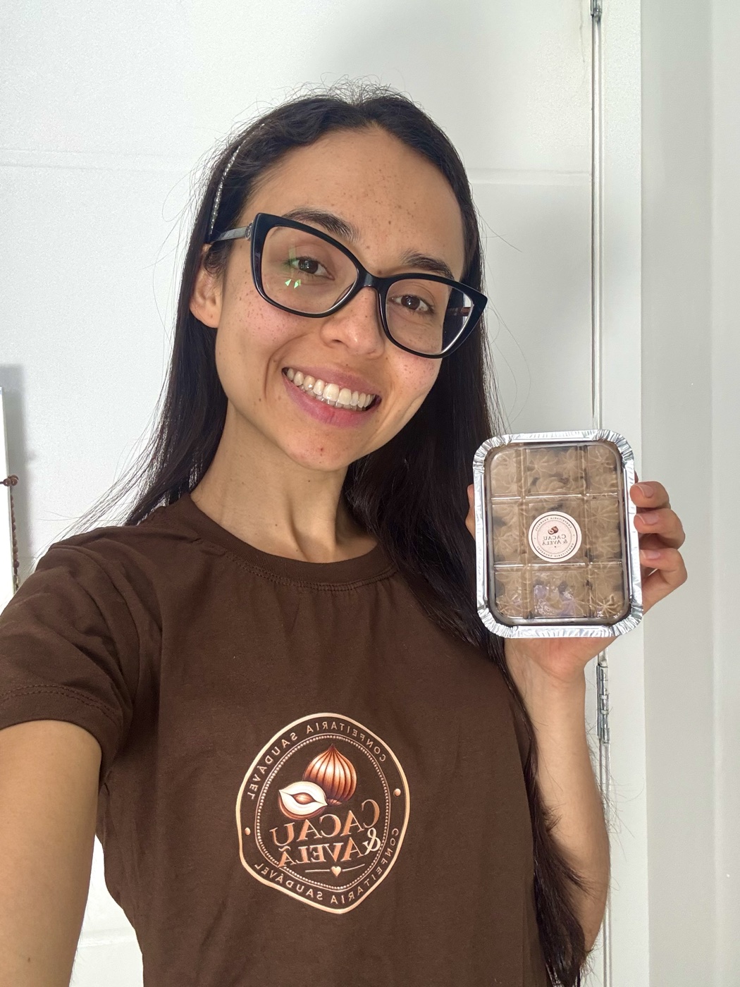 Gabi, de camiseta marrom com o selo da marca, segurando uma marmitinha de brownie